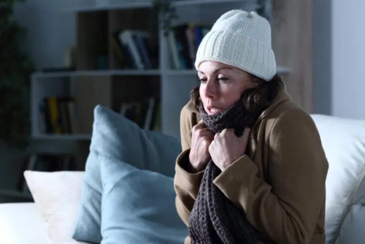 A woman sits on her couch, bundled in a hat and warm coat because she didn't prepare for a power outage.