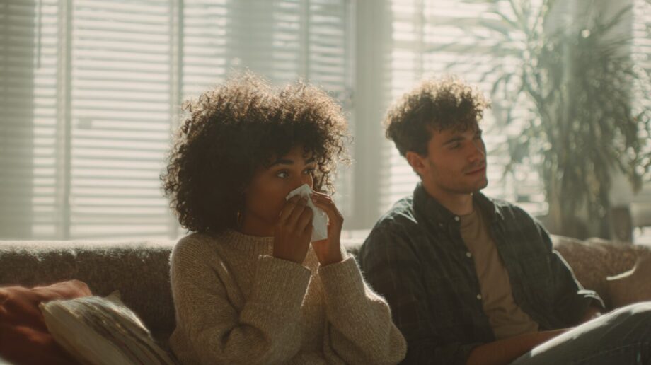 Couple in a Charlottesville, VA home relaxing on a couch during spring, with one person sneezing, suggesting indoor air quality issues from dust and pollen.